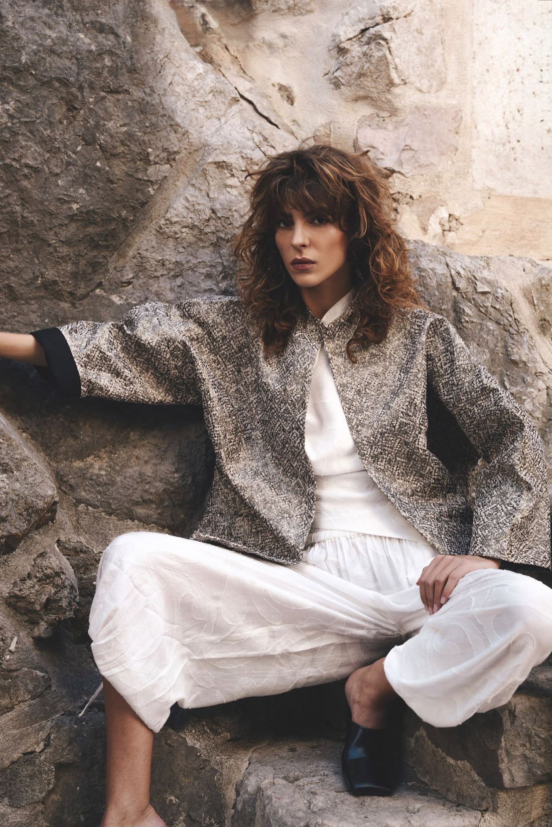Model in a textured oversized jacket and white trousers seated on stone steps